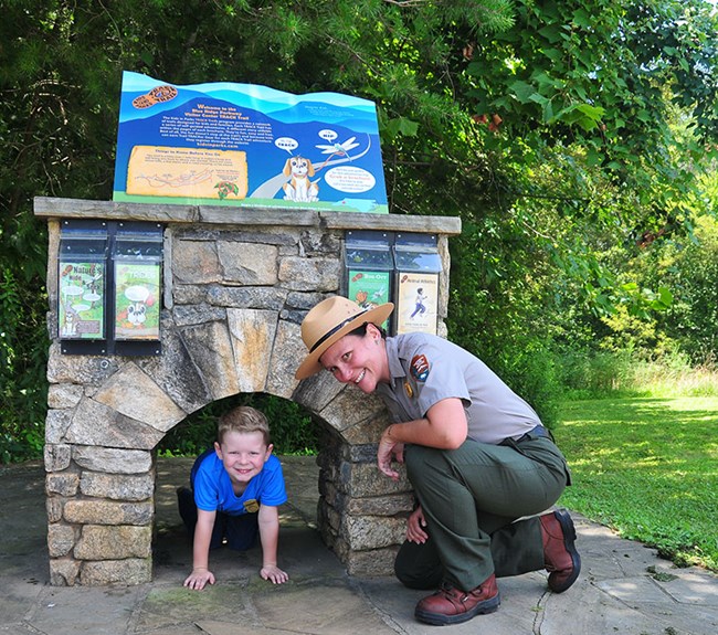 A young boy crawls through a small, stone arch while a kneeling, female ranger smiles.