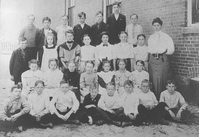 Group of young children sitting on the steps of schoolhouse. Black and white image