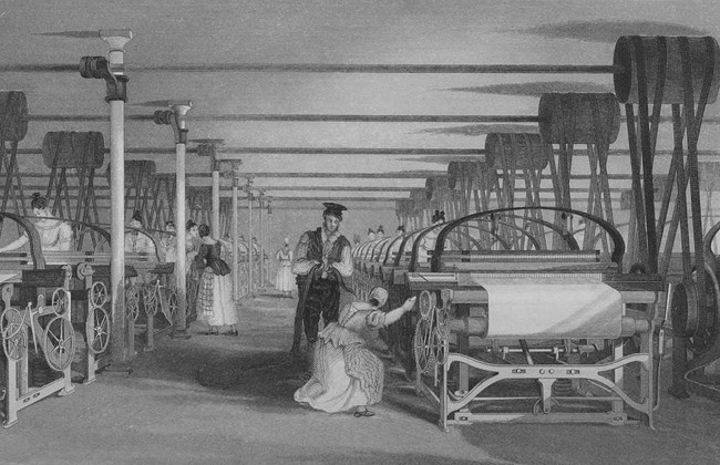 Male supervisor watches as female weavers work on power looms. Belts, wheels and large machines can be seen