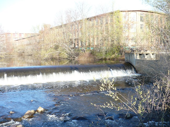 Dam with rolling water over the top of it. Whitin Machine Work factory building in the background