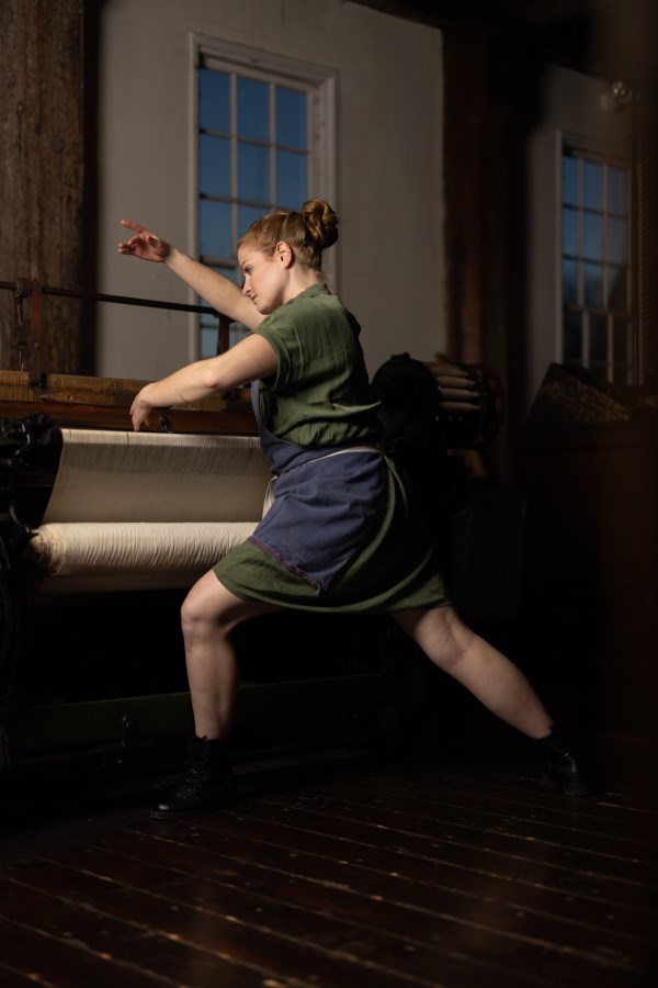 Women in green dress standing in front of loom in artistic pose with arms in the air