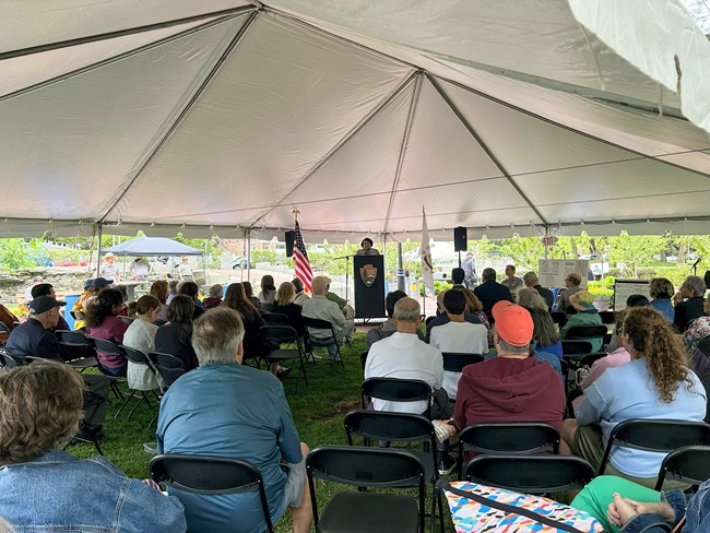 Group of people sit listening to Ranger talk. Ranger behind podium with NPS arrowhead