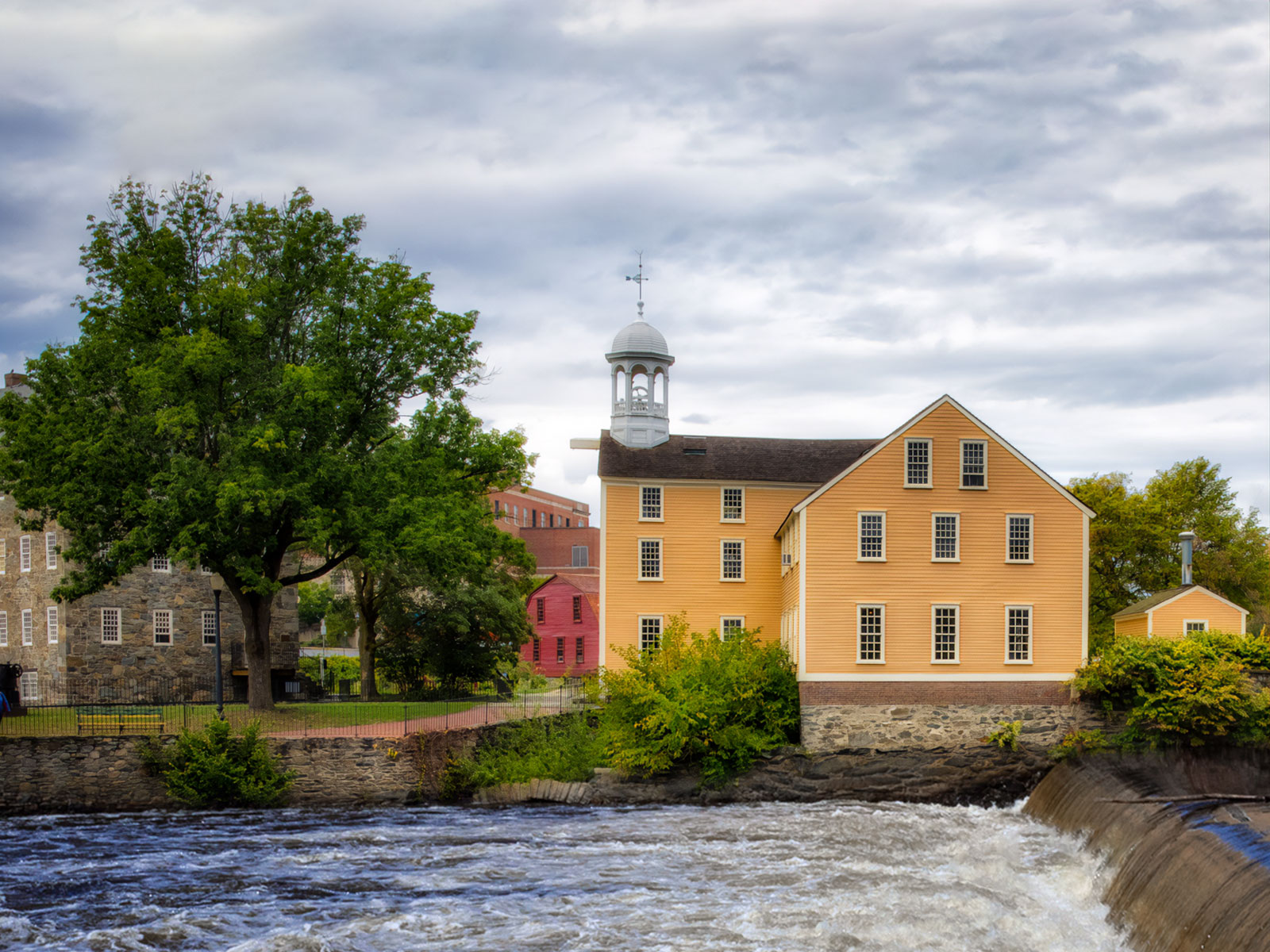 Cloudy day view looking across the river with the Wilkinson Mill building on the left the Sylvanus Brown House in the middle and the Slater Mill building on the right.