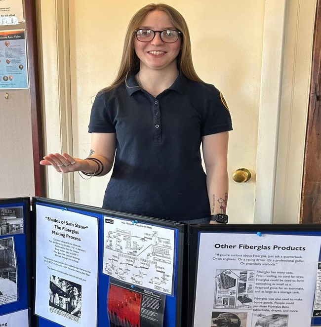 Emily Lady in blue polo wearing glasses stands behind a display board with information on it