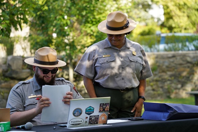 Man with beard wearing Ranger hat is seated next to standing woman in Ranger uniform