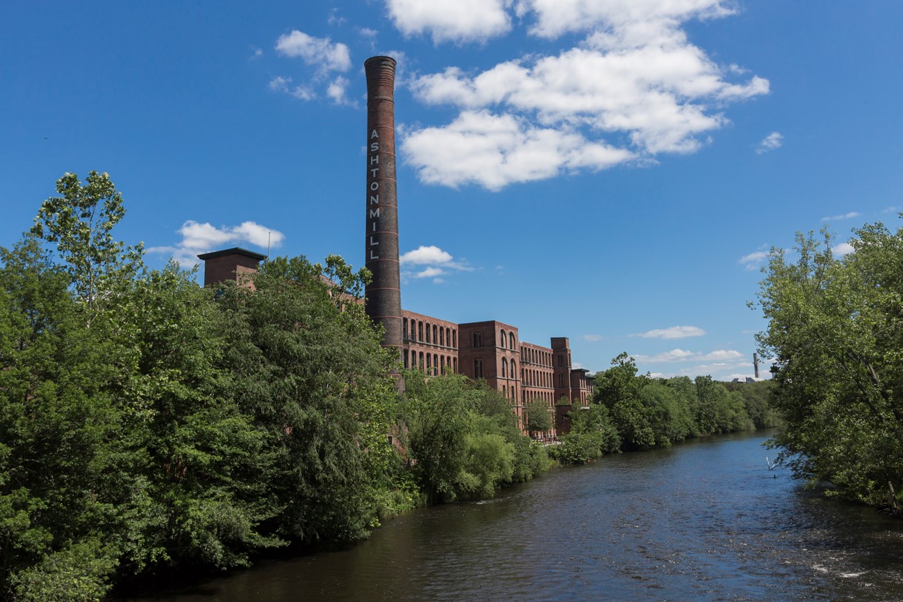 Large brick mill with smokestack along the Blackstone River