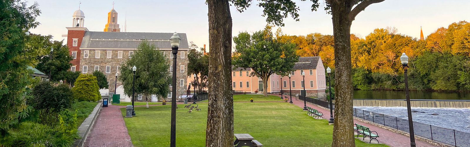 A tree-lined park with brick pathways, benches, and a river, with historic buildings in the background.