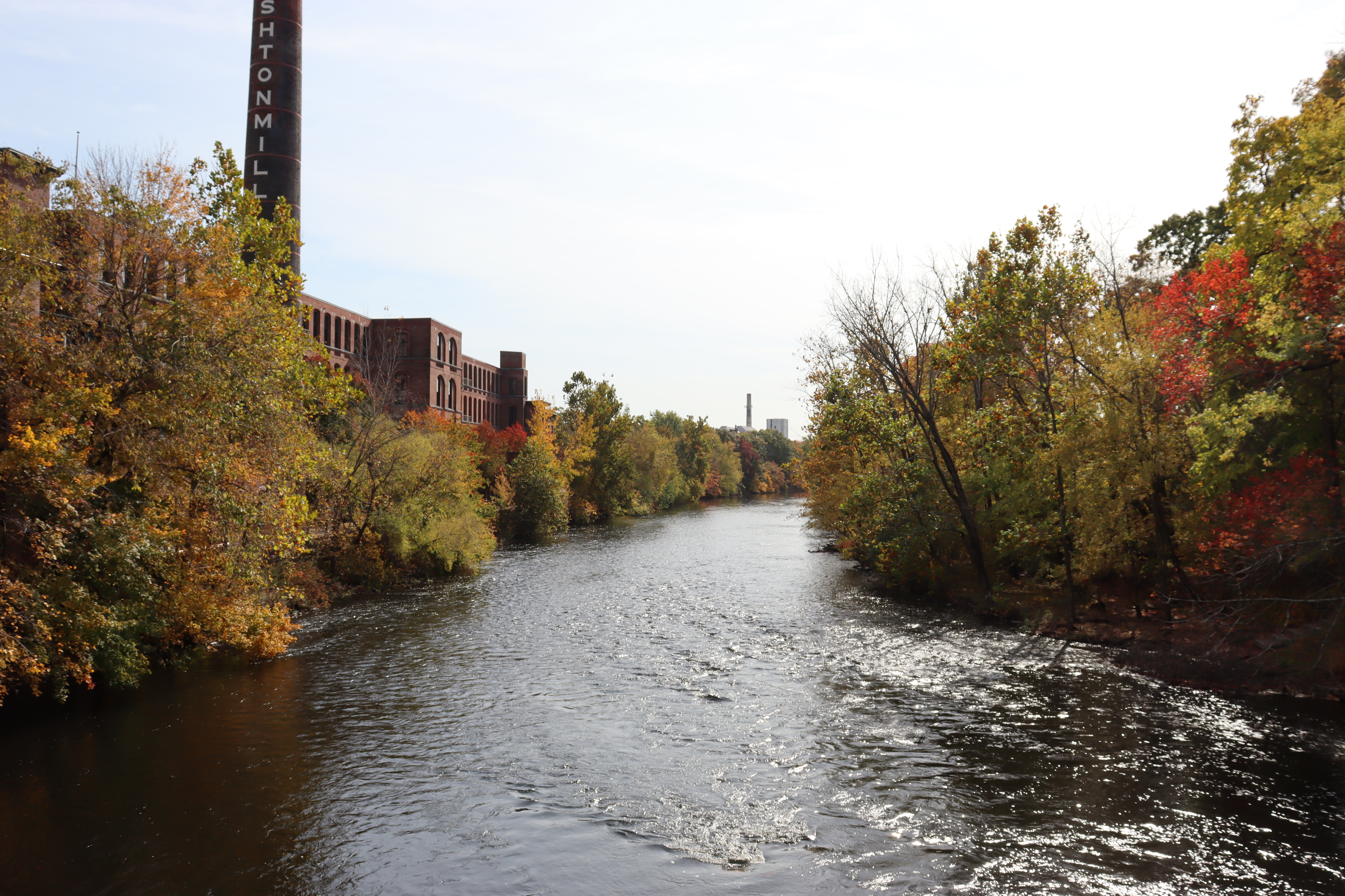 River with large brick mill and smokestack to the left bank. Trees surround the river with leaves colored yellow, brown, red, and green