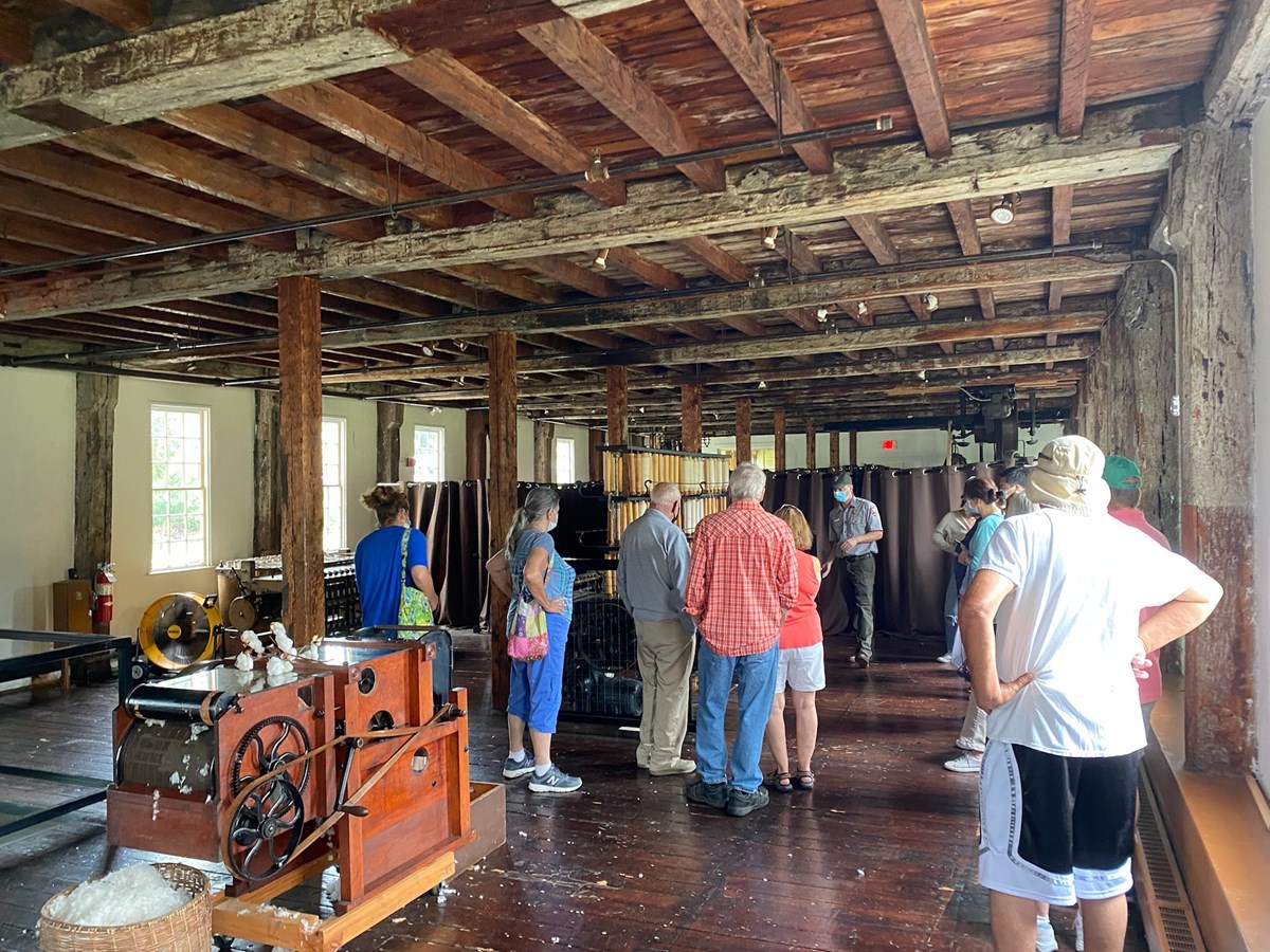 Park Ranger offers tour to large group of people inside Old Slater Mill. Machinery inside of an old building, visitors, and Park Ranger are visible.