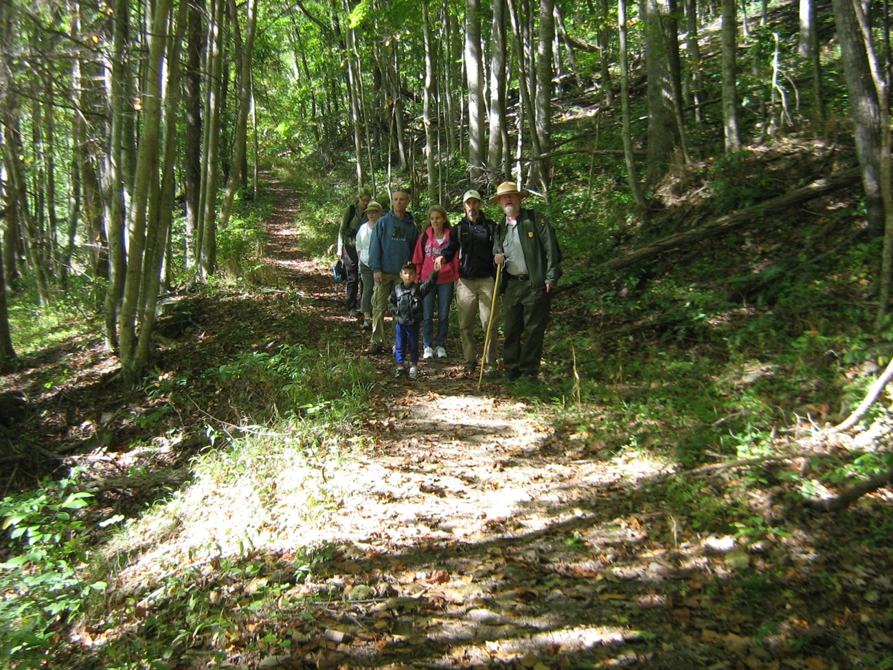 Photo of a National Park Ranger this is leading a guided hike on a tree lined trail with a group of visitors.
