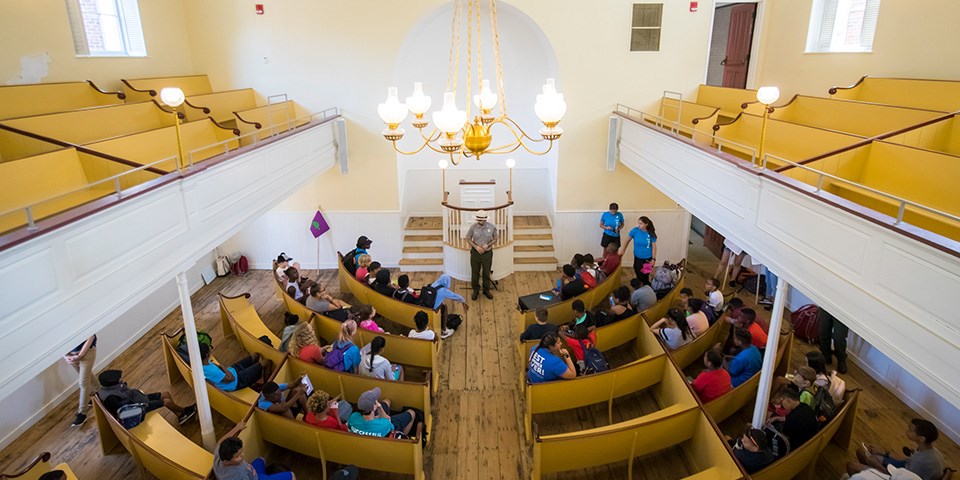 Looking down from a gallery, students sit in yellow arched pews listening to a park ranger speaking in front of a pulpit. Floors are knotted pine. A gold chandelier hangs overhead.