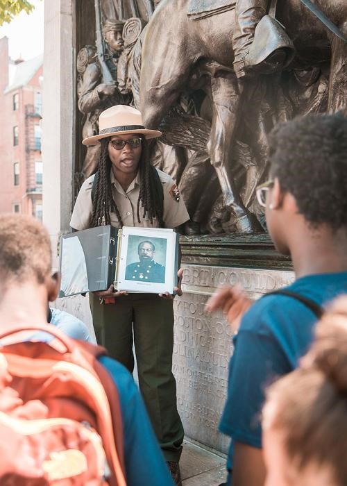 A park ranger in uniform and flat straw hat holds an open binder showing a photograph of a civil war soldier in a blue coat. Young students look in foreground. In background a bas relief sculpture made of bronze showing a horse and soldiers marching.