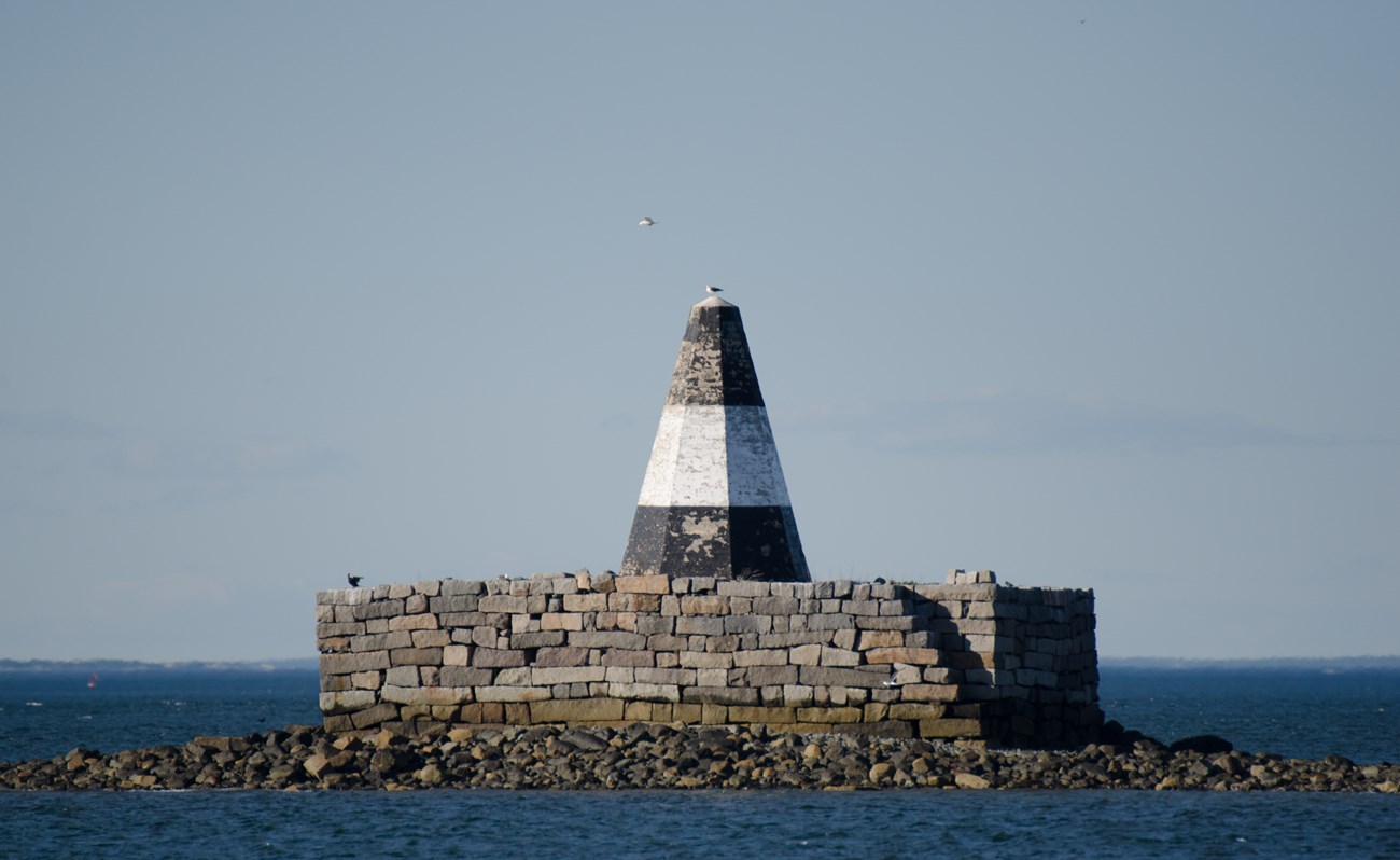 a white and black striped beacon that looks like a large traffic cone stands atop a brick base on a small island outcrop