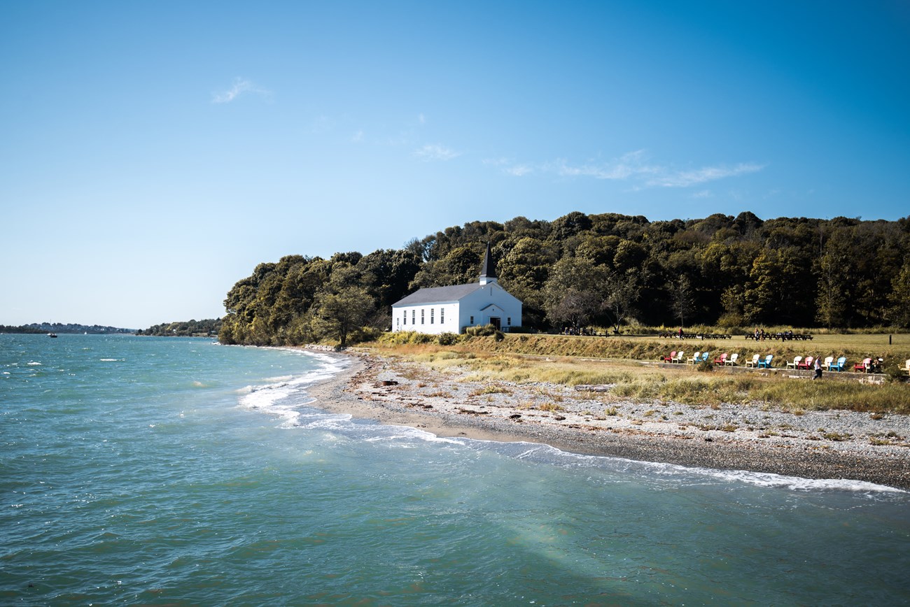 shoreline with low grass looking onto a field and wooded area. A white meeting house structure sits before the wooded area.