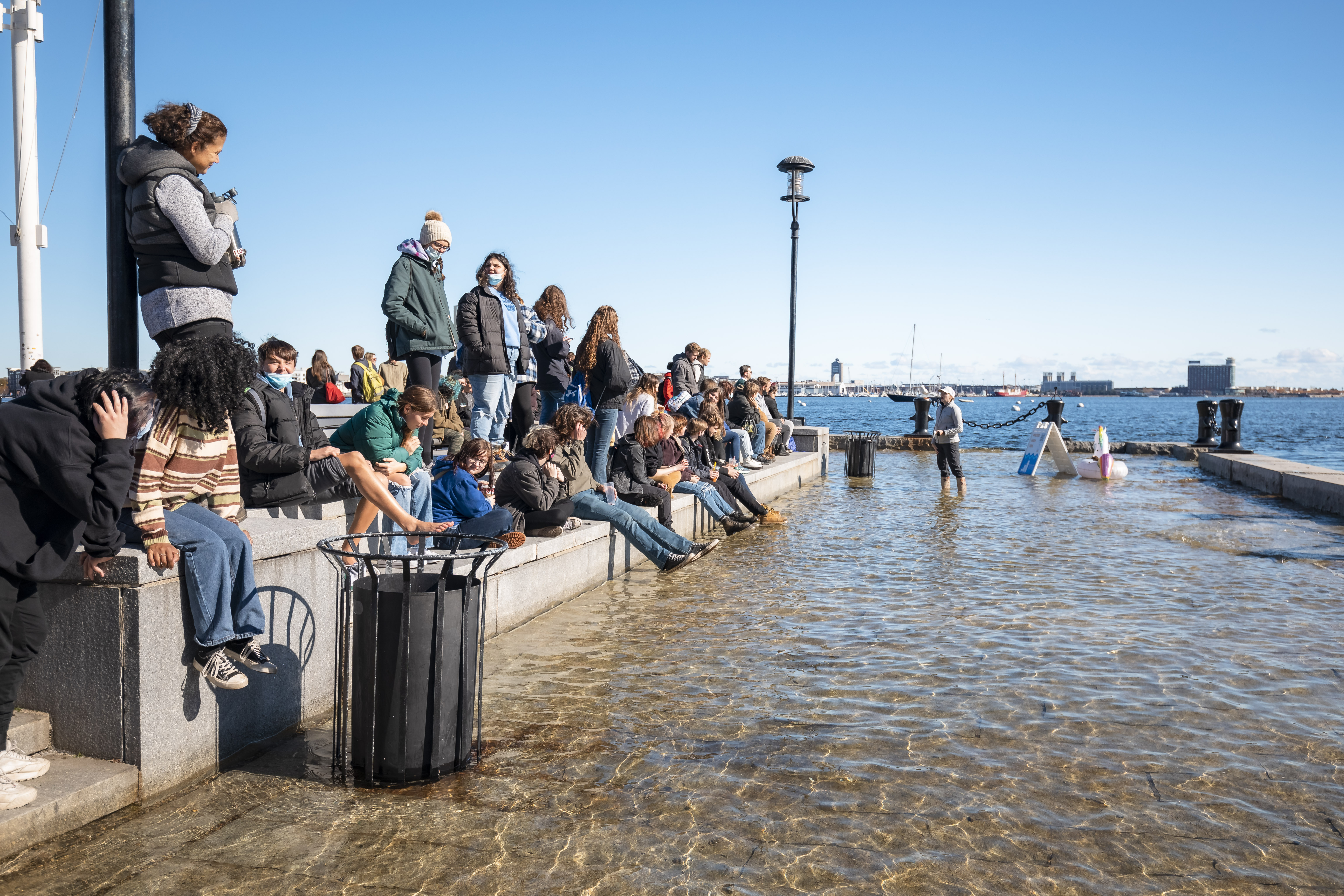 People sit along the harbor while someone speaks standing in water flooding the wharf