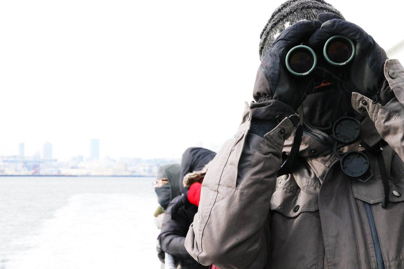 person in heavy coat and winter gear looking at the camera through binoculars