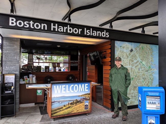 A park ranger stands in front of the Boston Harbor Islands Welcome Center. A sign that reads "Boston Harbor Islands" and another reads "Welcome." There are information materials and displays.