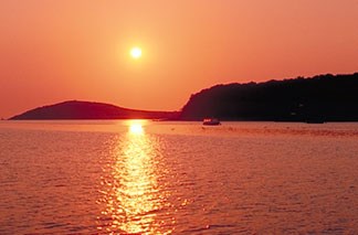 The sun sets over the Sheep Island. The bright orange of the sky and the yellow of the sun reflect off of the surrounding ocean. 