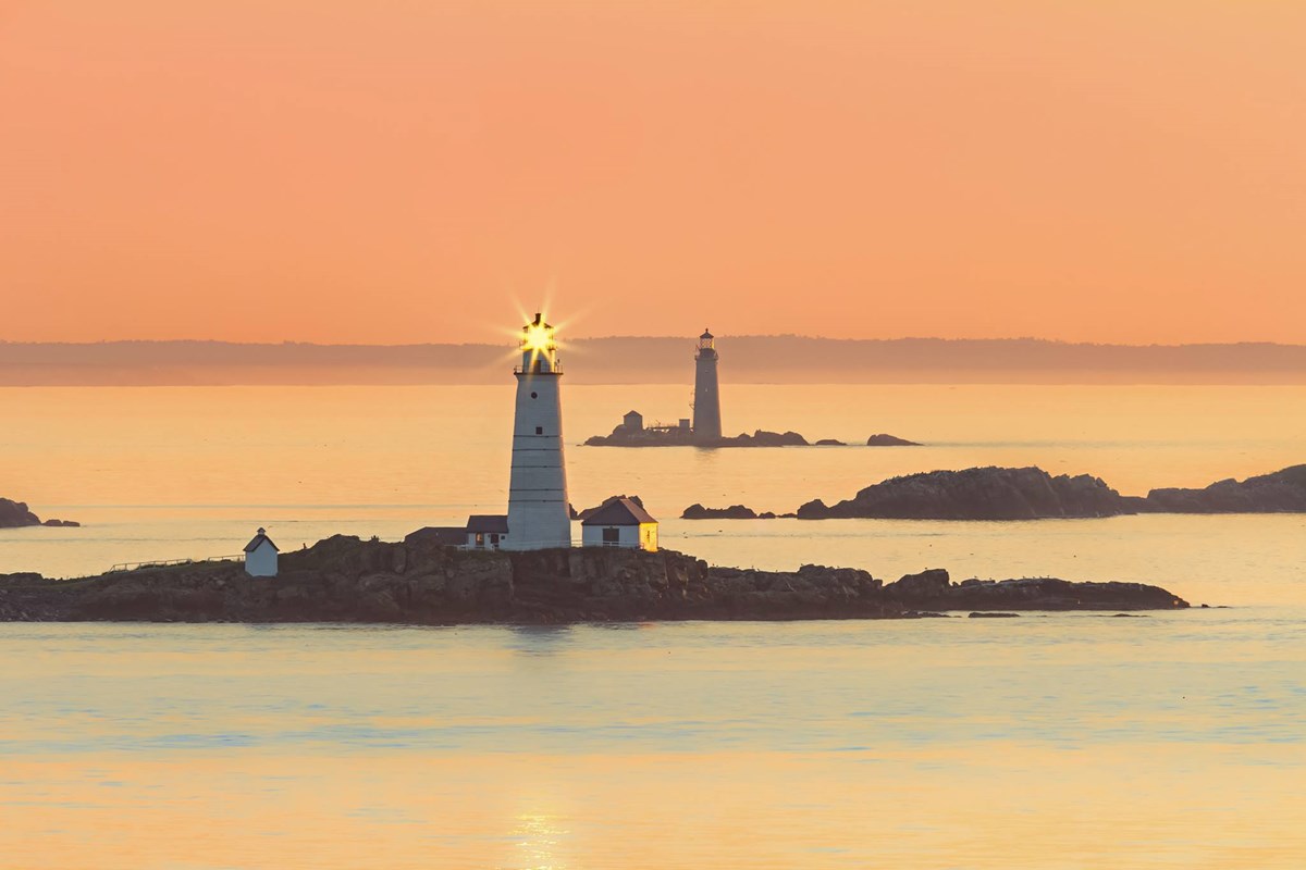 Lighthouse and small building on rocky island at sunset. Orange tinted sky reflected on still water.