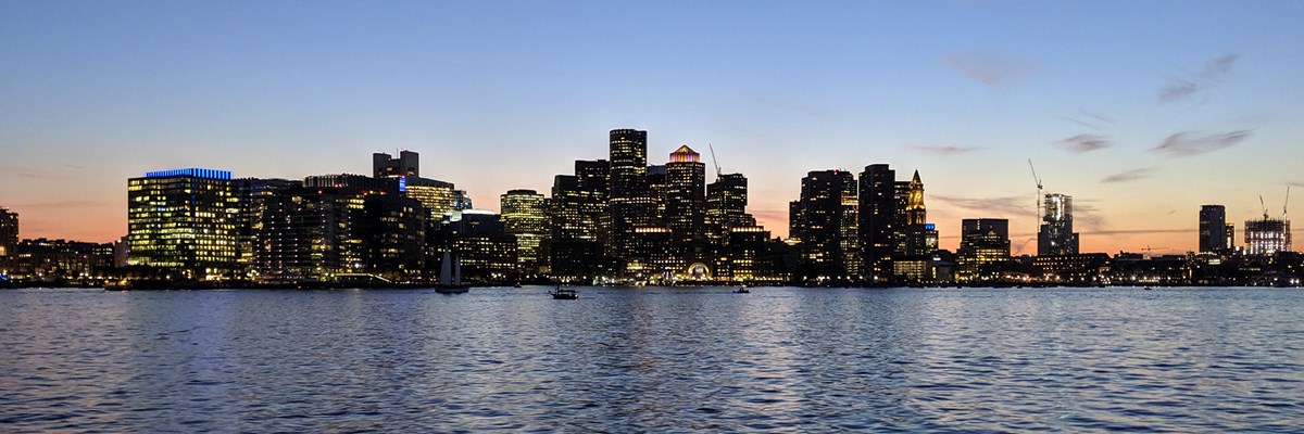 Wide view of Boston city skyline in the darkening light of sunset. View is from water looking at many towers with illuminated windows.