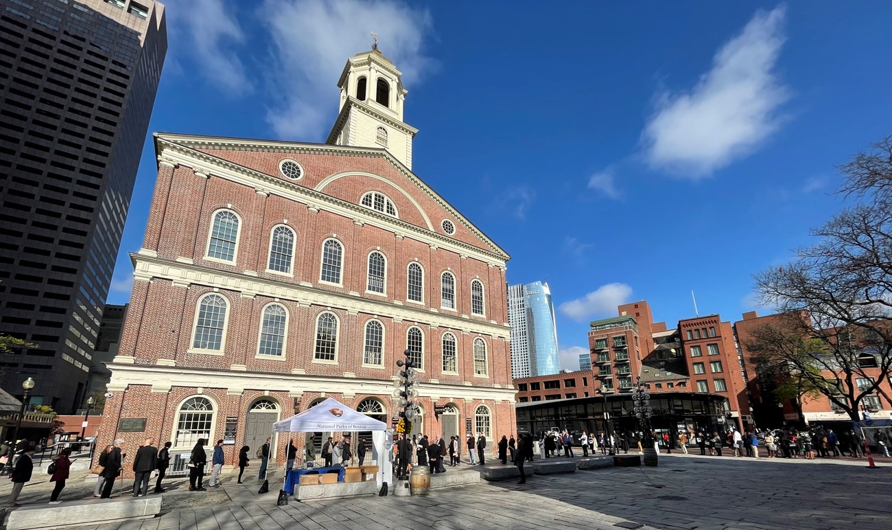 Faneuil Hall stands in the sunlight against a bright blue sky, an NPS pop-up tent is in front of it.