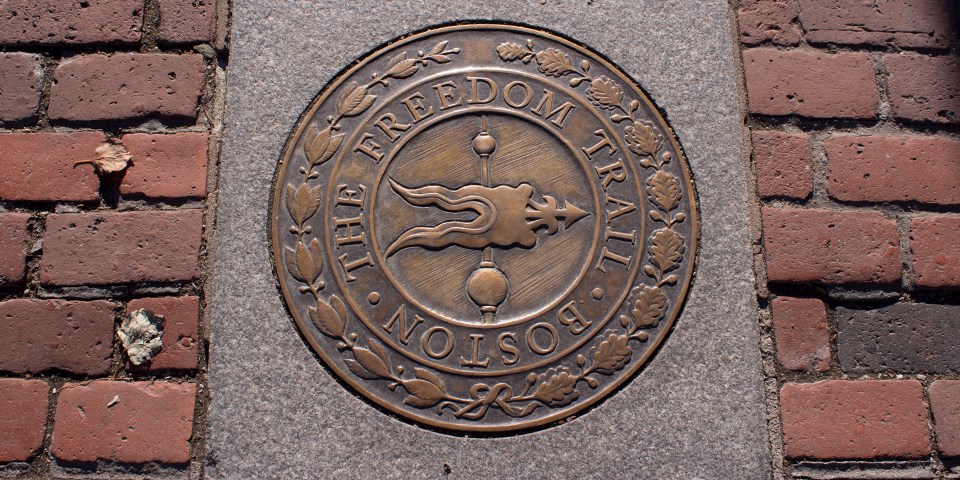 Bronze sidewalk marker medallion with words The Freedom Trail Boston. Weathervane bas relief is in center.