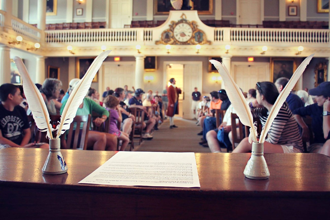 Photograph looking at a large assembly of seated people in an auditorium. In foreground are quill pens in inkwells at a table flanking a document. In background is a clock on a balcony.