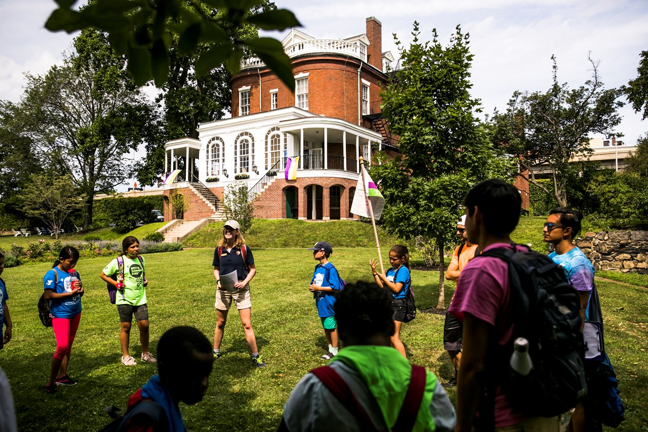 students in a big circle with education team member on grassy lawn in front of Commandant's House