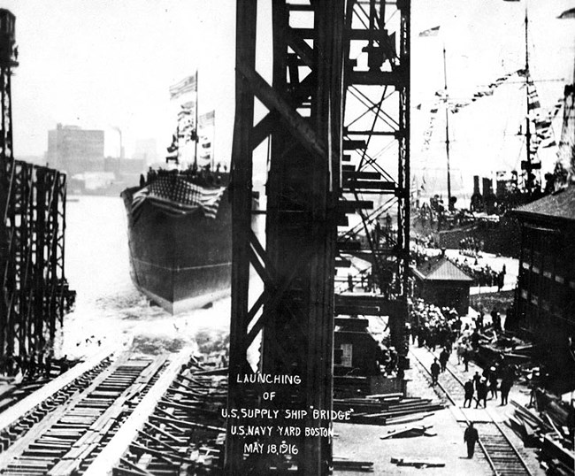 Photograph of a large supply ship sliding off the shipways into Boston Harbor.  Crowds are gathered on the pier to the right of the shipways, as both the supply ship and  USS Constitution to the right are dressed in flags.