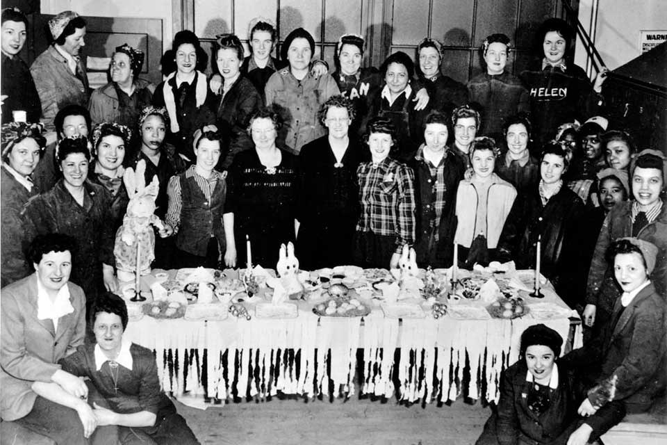 Black and white photograph of women shipyard workers around a table. Women are wearing working clothes and posing for the picture.