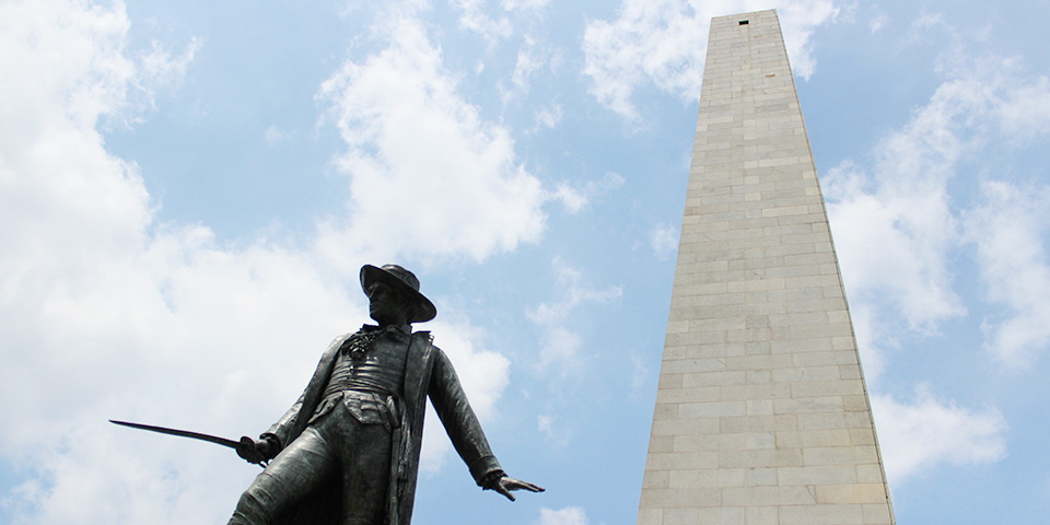 Photograph of the granite Bunker Hill monument with the bronze statue of William Prescott carrying a sword in the foreground