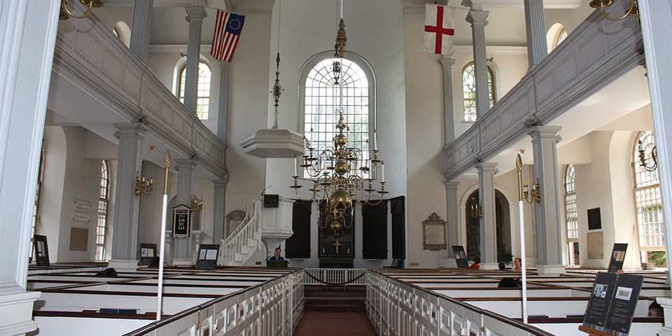Interior view of the church painted white with arched windows and many decorations. Rows of box pews cover the main floor. A speakers platform is on the front-left and a balcony supported by square columns is above.