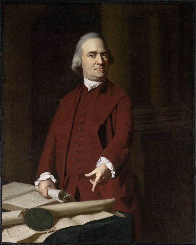 Adams standing in a red colonial suit pointing to a document on a table set before him.