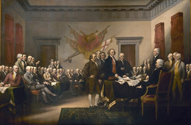 47 men in colonial suits sit or stand in a legislative chamber. In the foreground the five man committee who drafted the Declaration of Independence present their document to the seated President of the Congress John Hancock at the right of the painting.