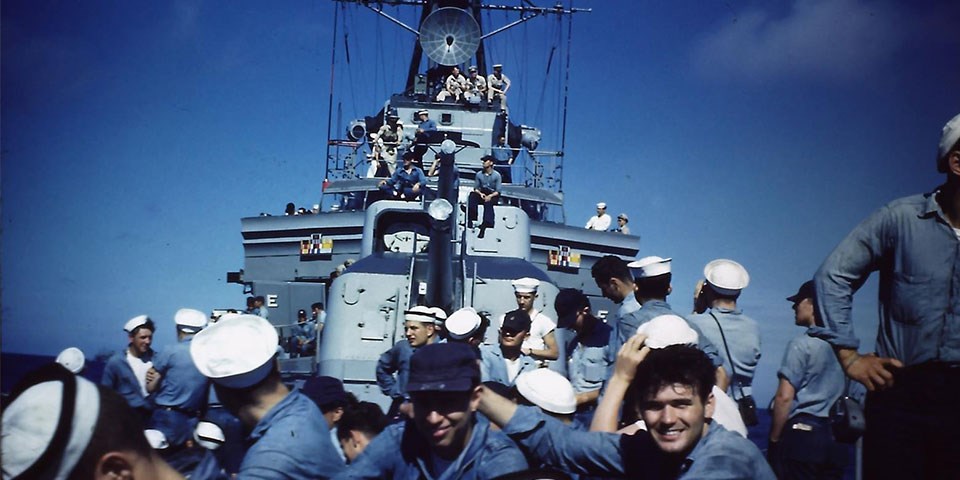 Photograph from the bow of Cassin Young looking aft with crewmembers on the deck, guns, and bridge. Crew are wearing dungarees and a few officers are wearing khaki uniforms.