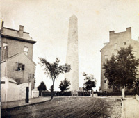 bh200.jpg Black and white photograph looking up a dirt road to a tall granite obelisk. Homes flank either side of the view up road.