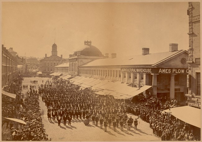 Yellowed photograph of a large parade of men next to long market buildings with shop names.