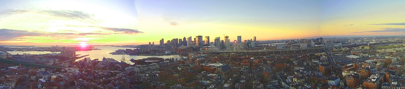 Blue skies with puffy white clouds over a downtown area with tall glass skyscrapers on a harbor waterfront. Horizon is orange as sun rises to the center left.