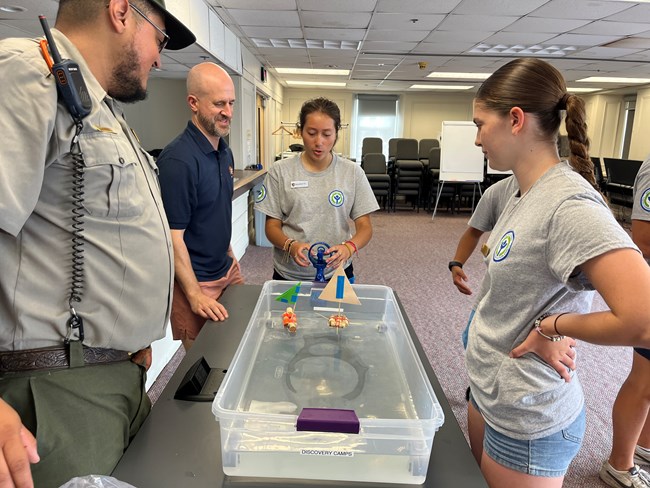 Two youth in a gray tshirt uniforms and two rangers stand at a table overseeing an sailboat activity