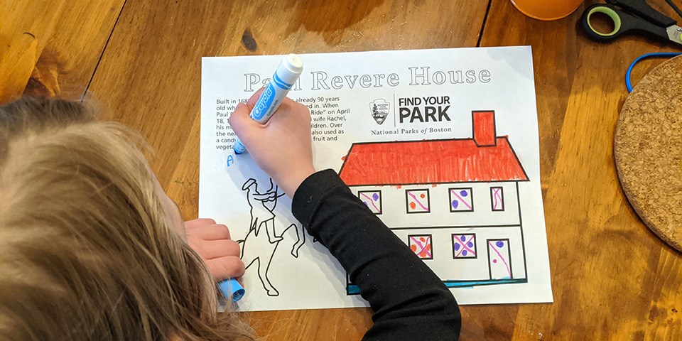 Child at table with marker coloring in a coloring sheet. Sheet reads "Paul Revere House" and features a two story house and a man on a rearing horse.