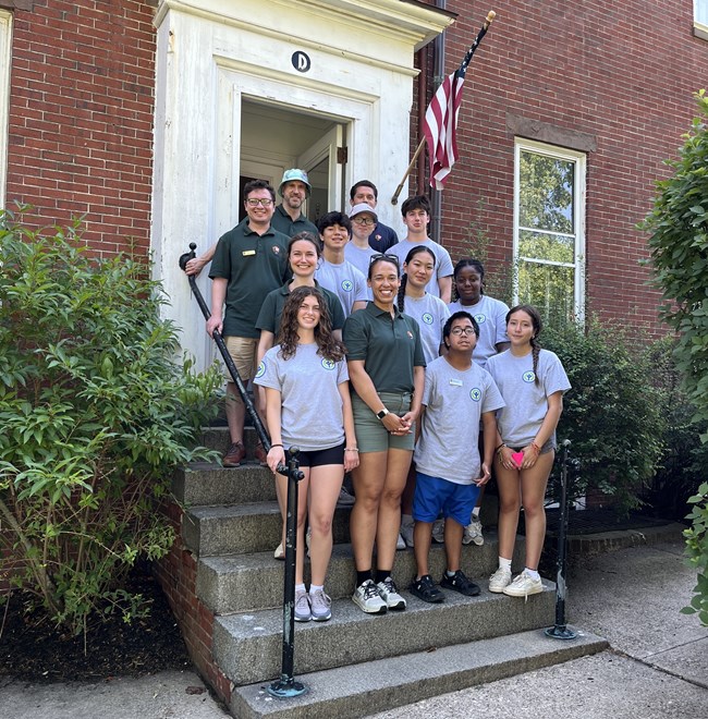 Multiple rows of youth stand in uniform gray tshirts in front of a brick building's entrance.