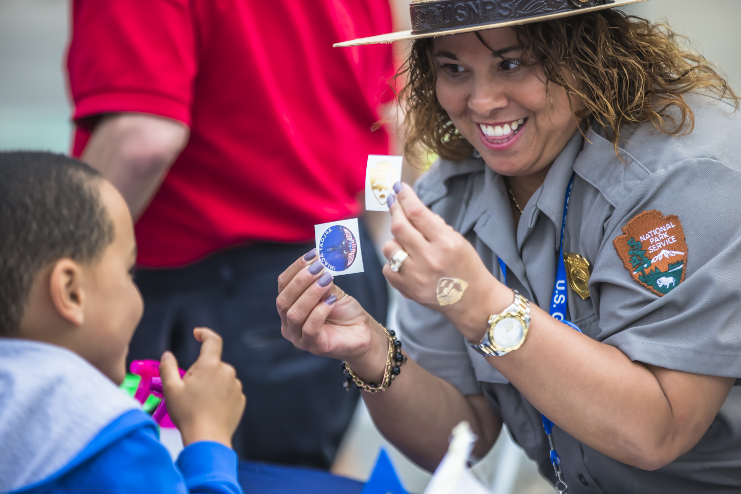 Ranger Veronica interacts with Young Visitors A female park ranger holds up a temporary tattoo and a sticker for a young visitor to choose from.