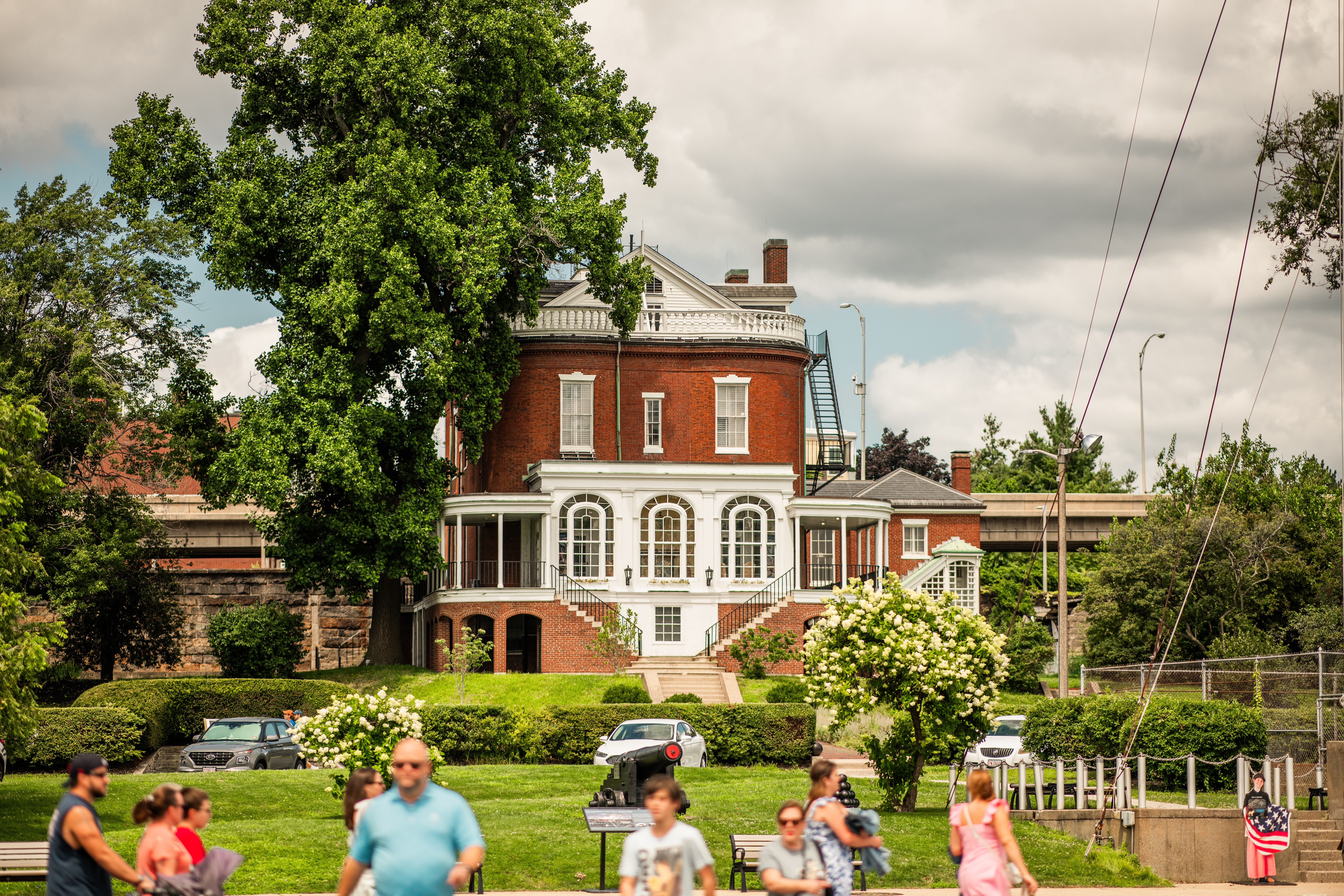 CommandantsHouse people are walking around the Charlestown Navy Yard. The Commandant's House, a federal-style brick building, sits on a lawn in the background.