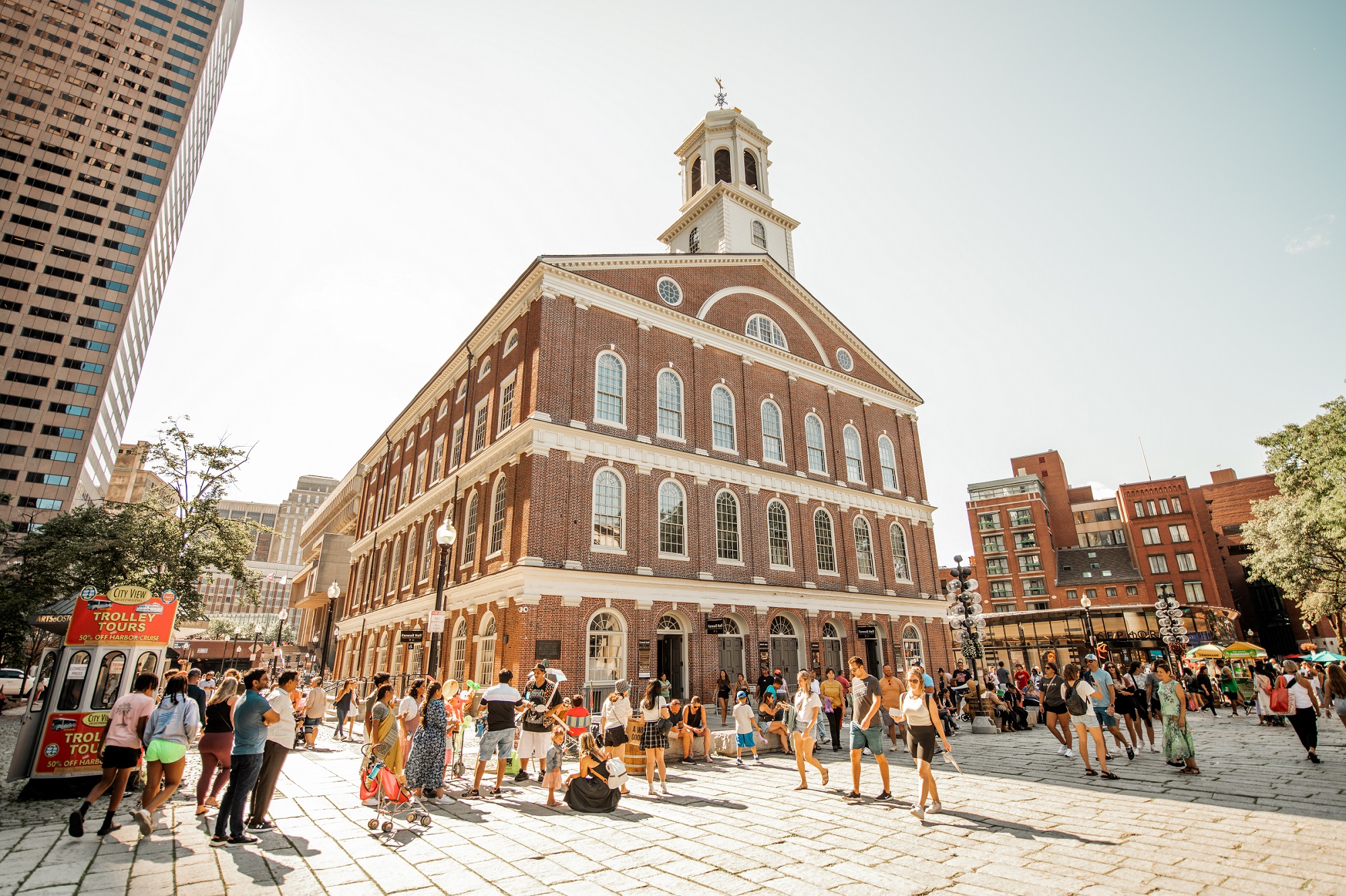 FreedomTrail-439-smaller Faneuil Hall, a red brick 3 story building with a cupola. Groups of people gather and walk around the square in front of the building.