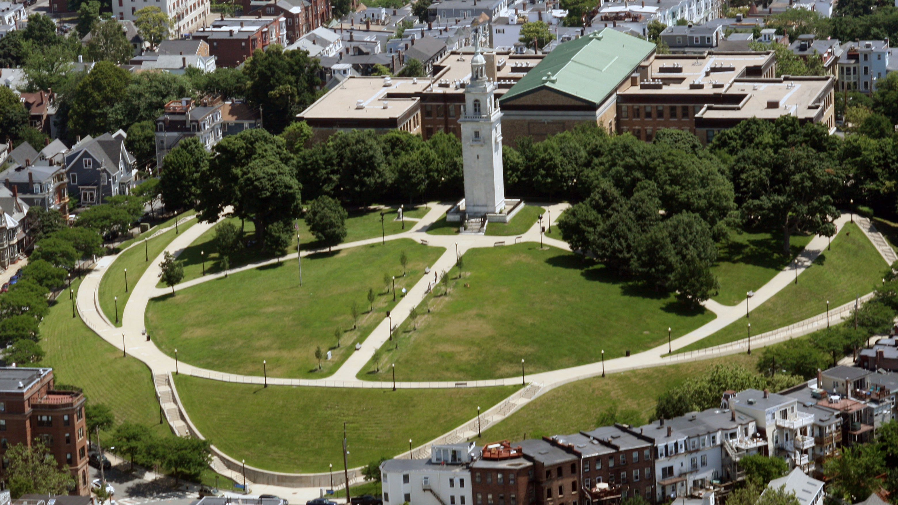Aerial photograph showing an elliptical park with grassy areas in the front and trees in the back. In between the trees stands a marble monument with square base, small cupola on top, with a spire.