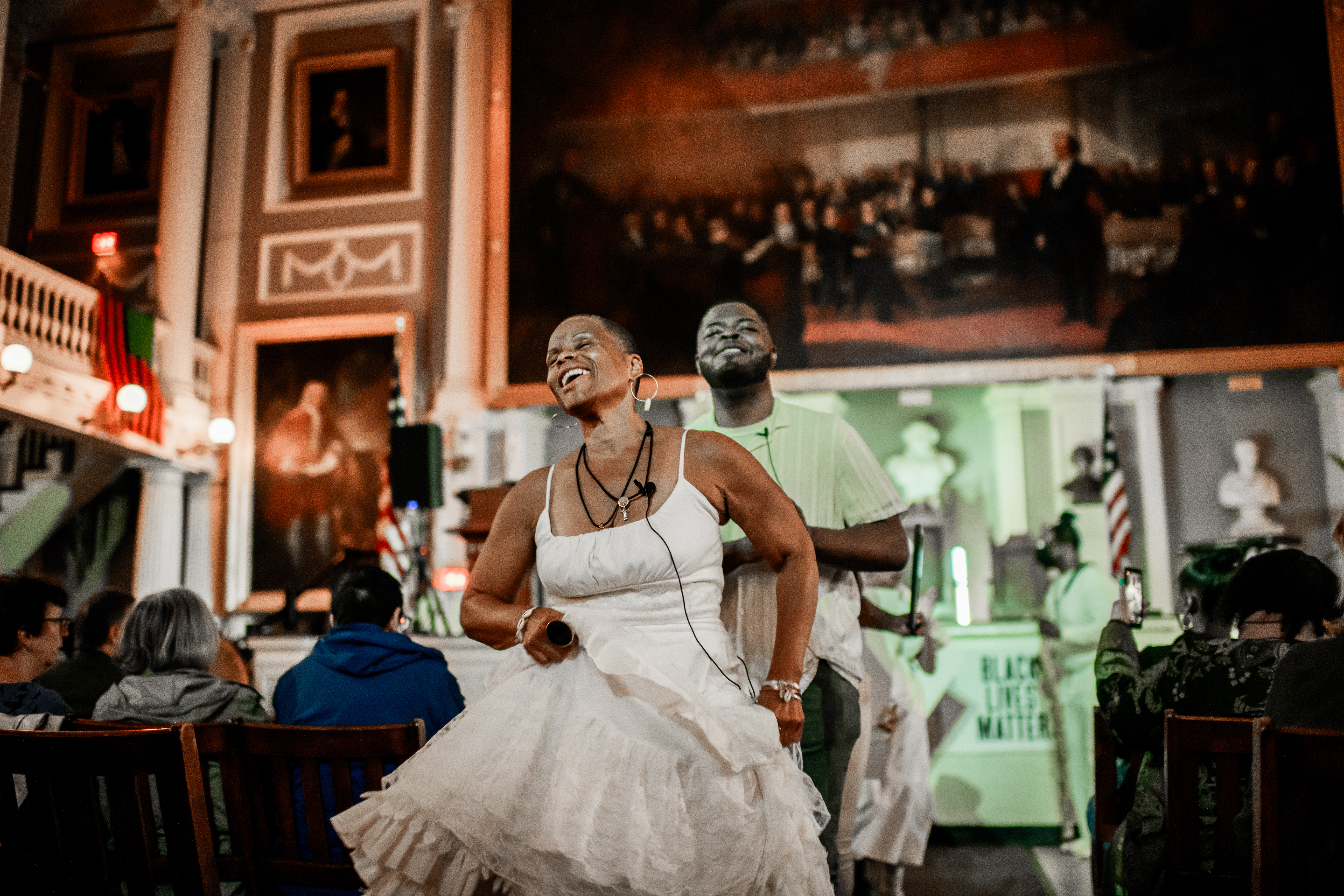 Photo by Arlan Fonseca. Courtesy National Park Service-National Parks of Boston Black woman in a white dress smiling and dancing as she leads a procession in Faneuil Hall