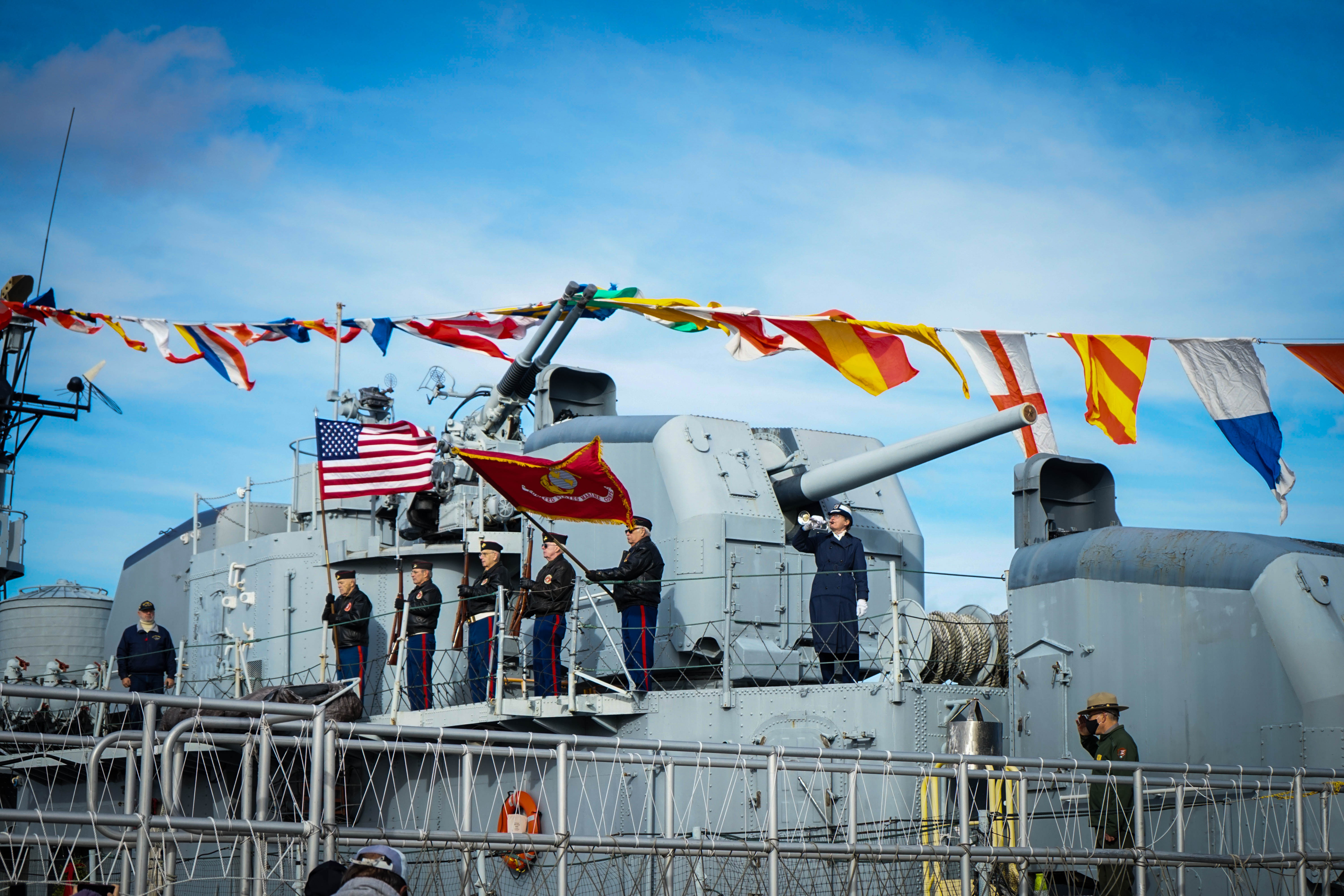 pearlharbor21-29-taps-JGould color guard with the US flag and US Marine Corps flag stands on an upper level of a naval ship dressed in pennants. A person plays on a bugle.