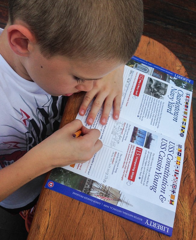 A young child works on a Junior Ranger book on a brown wooden table.