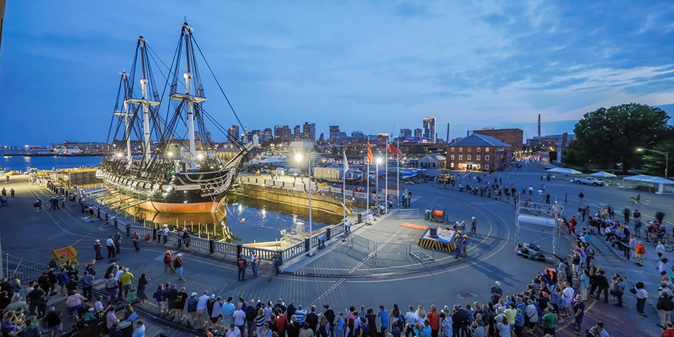 Wide view of a crowd on a pier. To left a three masted ship with shrouds floats in a flooded dry dock. Boston city skyline in background at twilight.