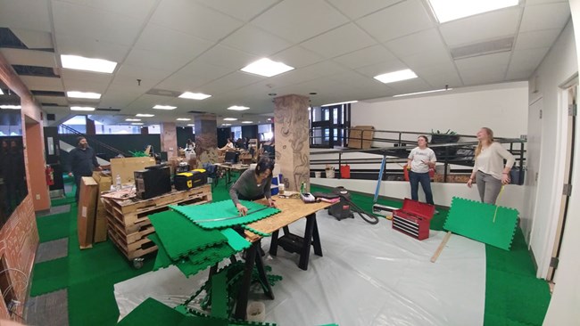 Photograph of park staff members measuring and cutting green carpet tiles in an open room.